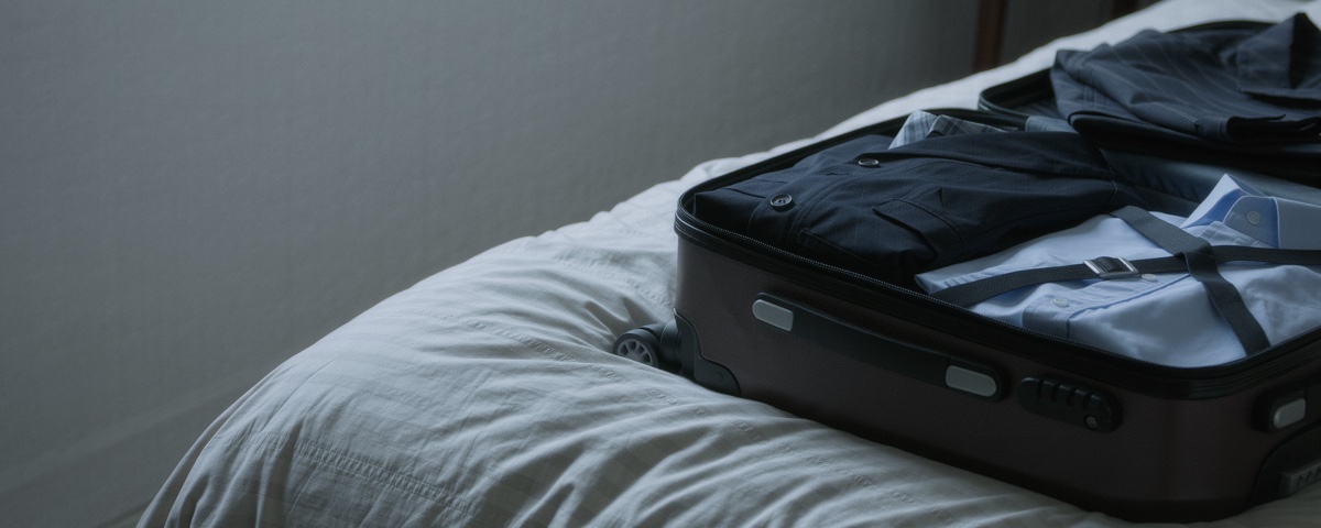 A half packed cabin case lying open on a made bed in a dim bedroom, folded shirts and a jacket inside it, plain bedding and bare wall to the left.