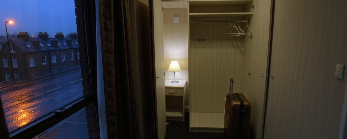 A small hotel room at dusk. On the left, a window looks out on a wet road and a brick terrace under an orange streetlight. On the right, a wardrobe stands open and completely empty except for bare hangers, with a closed suitcase upright on the floor and a single bedside lamp lit beyond it.