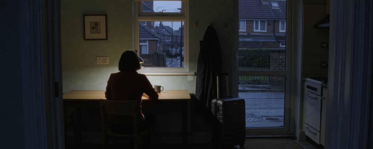 An ordinary kitchen before dawn. A woman sits at a small table to the left of centre with a mug in front of her, turned away from the camera, looking out at a grey wet street and the houses opposite. A coat hangs by the back door and a packed cabin case stands ready on the floor beside it.