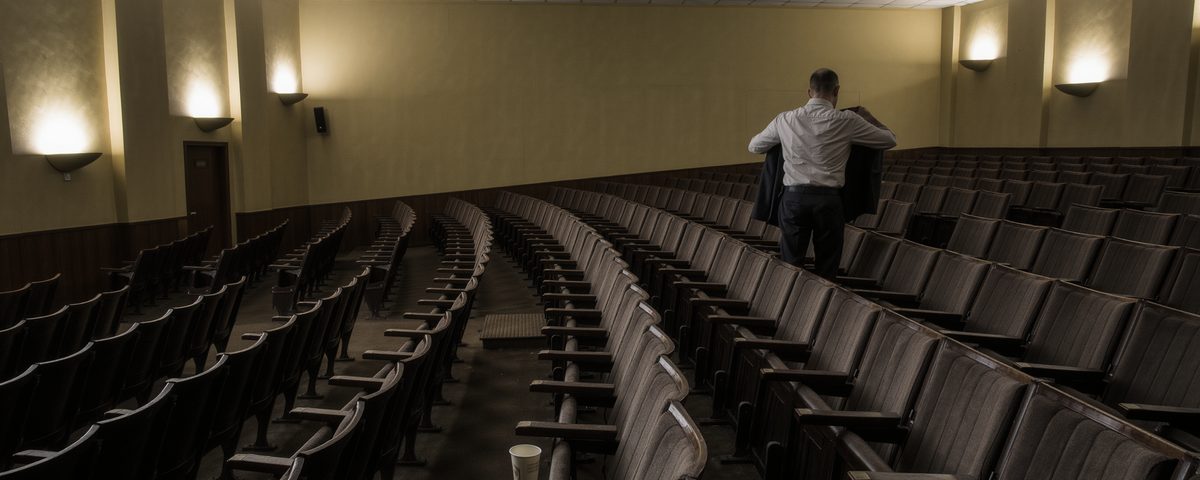 A man in a crumpled shirt pulling on his coat in the side aisle of an emptied tiered lecture room, rows of seats tipped up and the house lights brought half up.