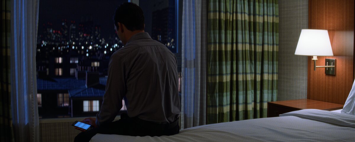 A hotel room at night. A man in a work shirt sits on the edge of the bed to the left of centre, turned away from the camera toward the window, a lit phone held loosely in his hand at his side. Rooftops and the scattered lights of the city fill the window. A lamp is on against the wall to the right, the bed beside him empty.