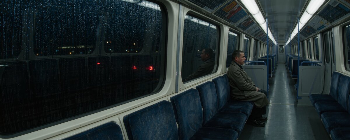 The inside of an almost empty train carriage at night. Rain runs down the black window on the left, red lights showing somewhere outside. One man in a work coat sits alone on the blue seats to the right of centre, turned away toward the glass, his reflection beside him, with the empty aisle running away behind him.
