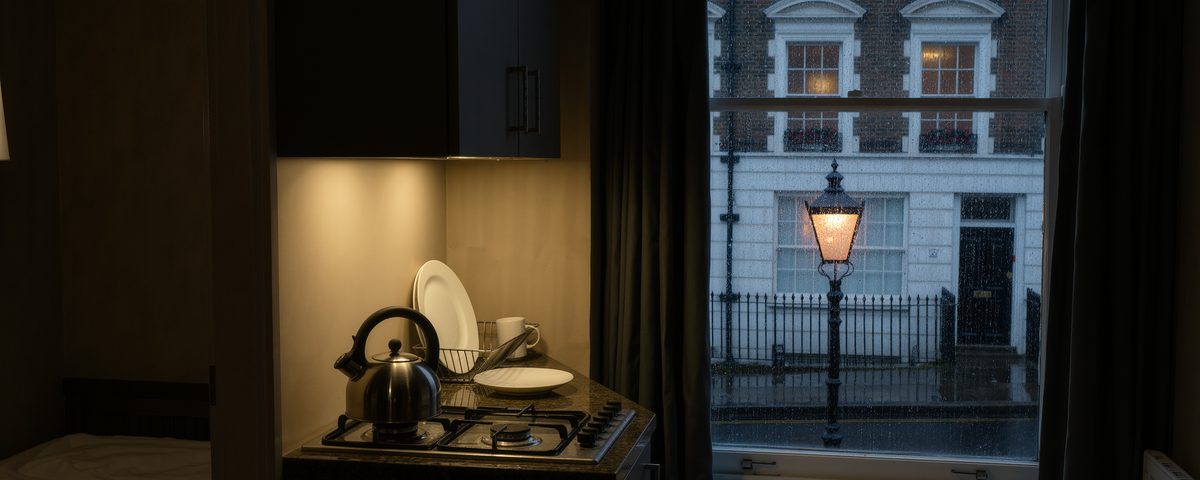 A small aparthotel kitchenette lit by a single under cupboard light, with a kettle, one plate and one mug on the drainer, looking out through a sash window at a wet London stucco terrace and a glowing street lamp.