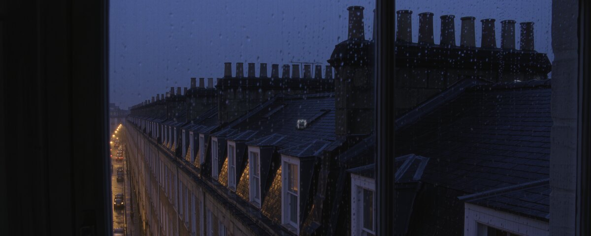 The view from an upstairs window on a rainy evening, rain beaded on the glass and the dark frame across the middle of the frame. Below and to the left, a terraced street falls away with its lamps already lit and cars parked along it. Slate roofs, dormer windows and rows of chimney pots run off toward a flat grey horizon.