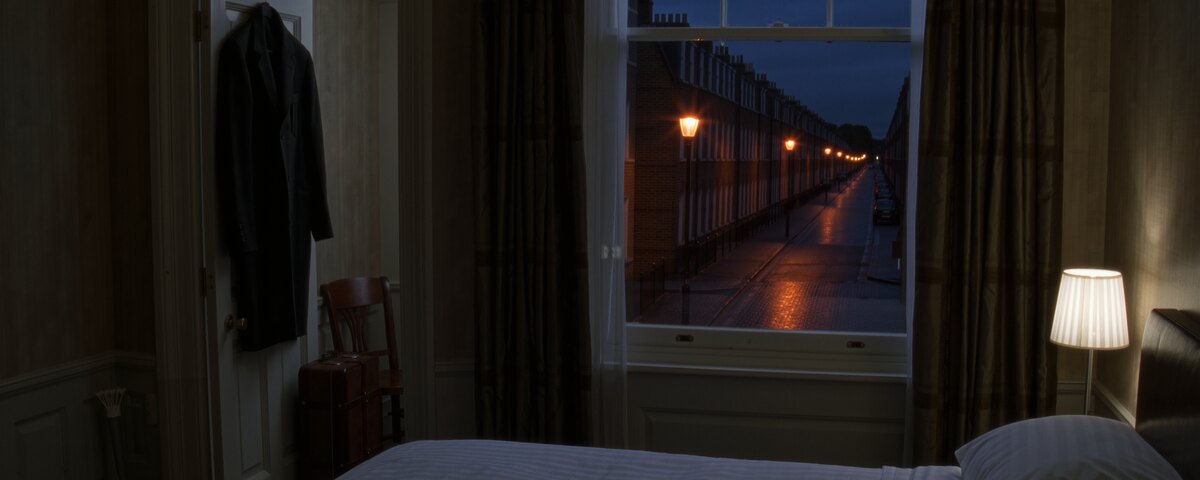 A hotel room in the evening with nobody in it. A coat still hangs on the back of the open door on the left, an unopened case on the floor beneath it. Through a tall sash window in the middle of the frame, a terraced street runs away under orange lamps, the road wet. The bed is made and a bedside lamp is lit on the right.