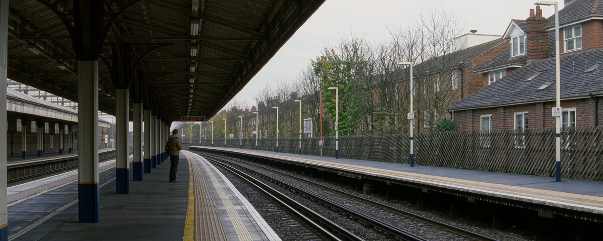 An empty suburban railway platform on an overcast morning, seen along its length under an old iron canopy. One person in a casual jacket with a small bag stands off to the left, turned away, looking down the track. The platform opposite is deserted, with bare trees and the backs of brick houses behind it.